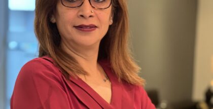 A woman wearing a red shirt and glasses, looks at the camera with folded hands