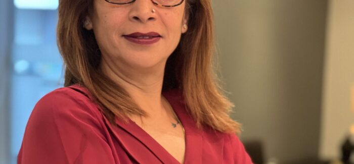 A woman wearing a red shirt and glasses, looks at the camera with folded hands