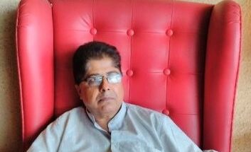 Image shows a man wearing a white kurta sitting on a large red chair