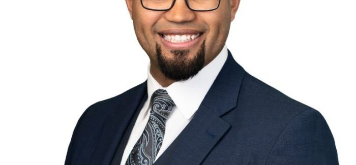 Image shows a bald man with glasses and a french beard, wearing a suit and smiling at the camera