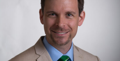 Image shows a man with a light french beard, wearing an off white suit, smiling at the camera