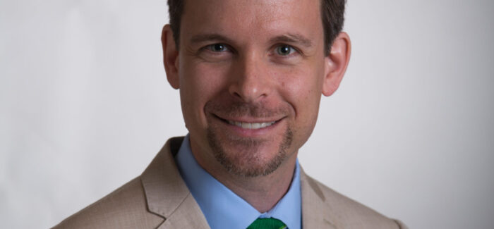 Image shows a man with a light french beard, wearing an off white suit, smiling at the camera