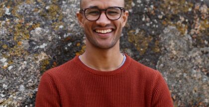 Image shows a bald man with glasses, and a red sweater smiling at the camera