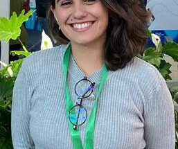 A woman wearing a grey shirt smiles at the camera. She has a green tag hanging around her neck.