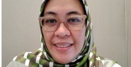 A woman wearing a green hijab and prescription glasses smiles at the camera